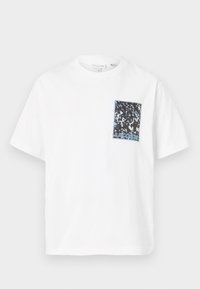 Camiseta blanca de manga corta con un bolsillo a rayas negro y azul en el lado izquierdo del pecho. La tela parece suave; diseño simple y sin adornos.