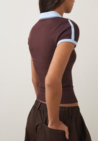 Vrouw draagt een nauwsluitend bruin kort shirt met een lichtblauwe kraag en streep, gecombineerd met een bruine broek, hand in achterzak.