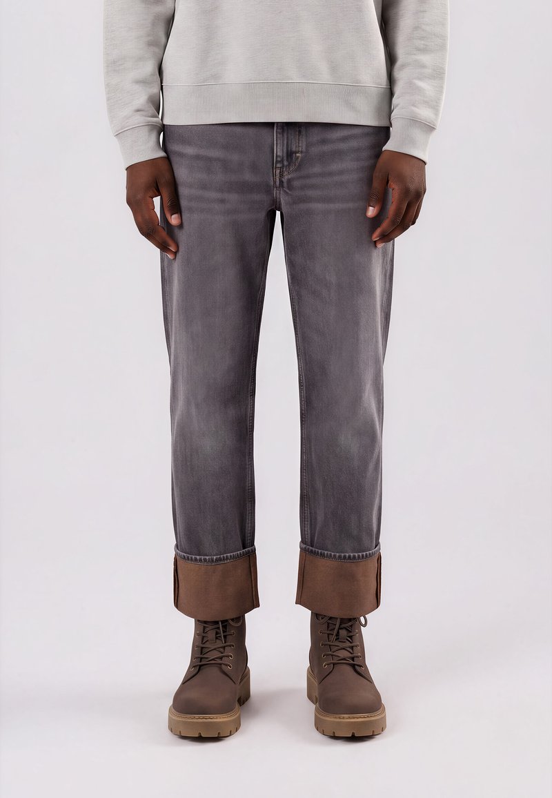 Personne portant un jean gris retroussé aux chevilles et des bottes marron à lacets avec semelles beiges, debout devant un fond uni.