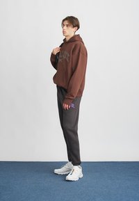Brun hette med forlommes og grafisk detaljering, kombinert med mørkegrå joggebukser med lilla logo. Hvite sneakers med fargerike aksenter.