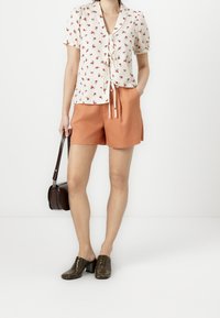Camisa corta de manga con estampado floral y un lazo en la cintura, combinada con pantalones cortos naranjas. La persona lleva mules de cuero marrón y porta una pequeña bolsa marrón.
