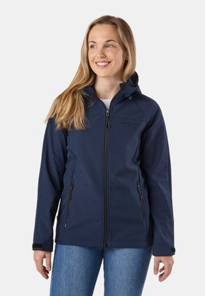Femme portant une veste à capuche zippée bleu marine avec poches zippées et un jean bleu, souriante et regardant vers la droite sur un fond uni.