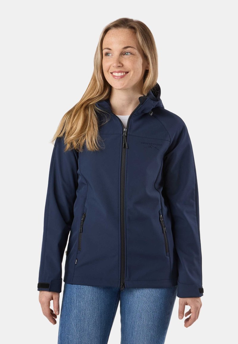 Femme portant une veste à capuche zippée bleu marine avec poches zippées et un jean bleu, souriante et regardant vers la droite sur un fond uni.