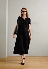 Black short-sleeve dress with a fitted bodice and flowing skirt. Model carries a large black tote bag and wears dark sunglasses with flat shoes.