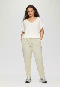 Weißes V-Ausschnitt-Oberteil und hellbeige Jogginghosen mit elastischem Bund und Kordelzug. Model trägt silberne Turnschuhe, lange lockige Haare sind sichtbar.