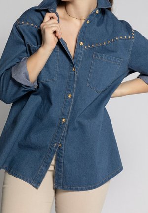 Kvinde iført stor blå denimskjorte med opkrullede ærmer, guldknapper og guldfarvede nitter på tværs af brystet, kombineret med beige bukser.