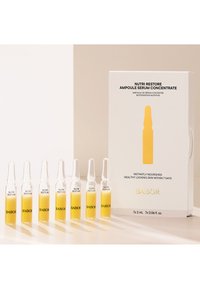 Sept petites ampoules en verre contenant un sérum jaune étiqueté "BABOR Nutri Restore" à côté d'une boîte blanche de produit avec un marquage similaire.