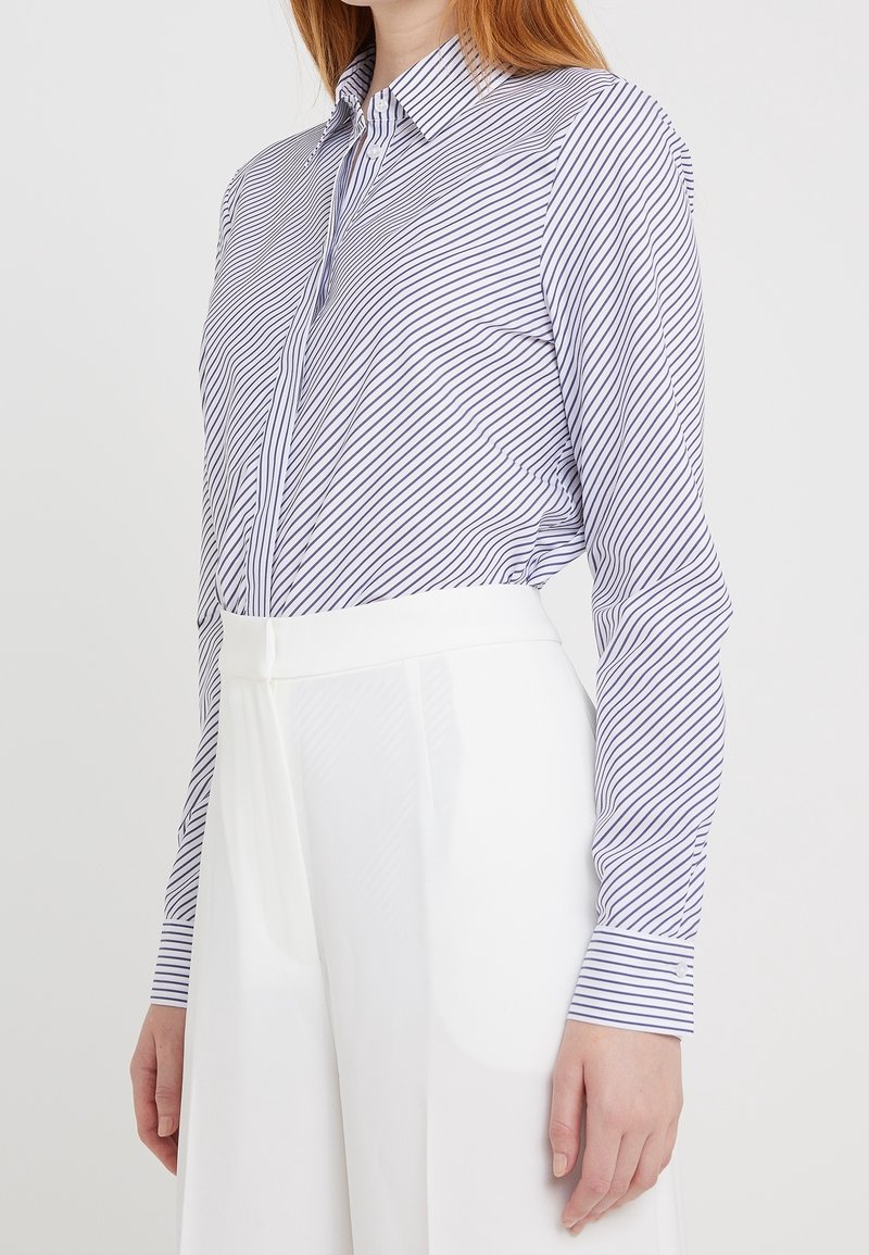 Femme portant une chemise blanche à manches longues avec de fines rayures noires, rentrée dans un pantalon blanc taille haute à jambes larges.