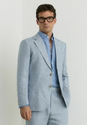 Mees, kes kannab musti prille, helesinist jakk-särki, sobivaid pükse ja sinist nööbitavat särki, seisab ühevärvilise valge seina taustal.