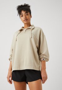 Felpa beige con collo a mezza zip, vestibilità comoda e polsini a costine. Include un piccolo logo sulla manica sinistra. Pantaloni corti neri.