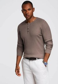 Camisa henley marrón de manga larga y canalé con tres botones negros en el cuello, combinada con pantalones claros. Se ve un reloj plateado en la muñeca.