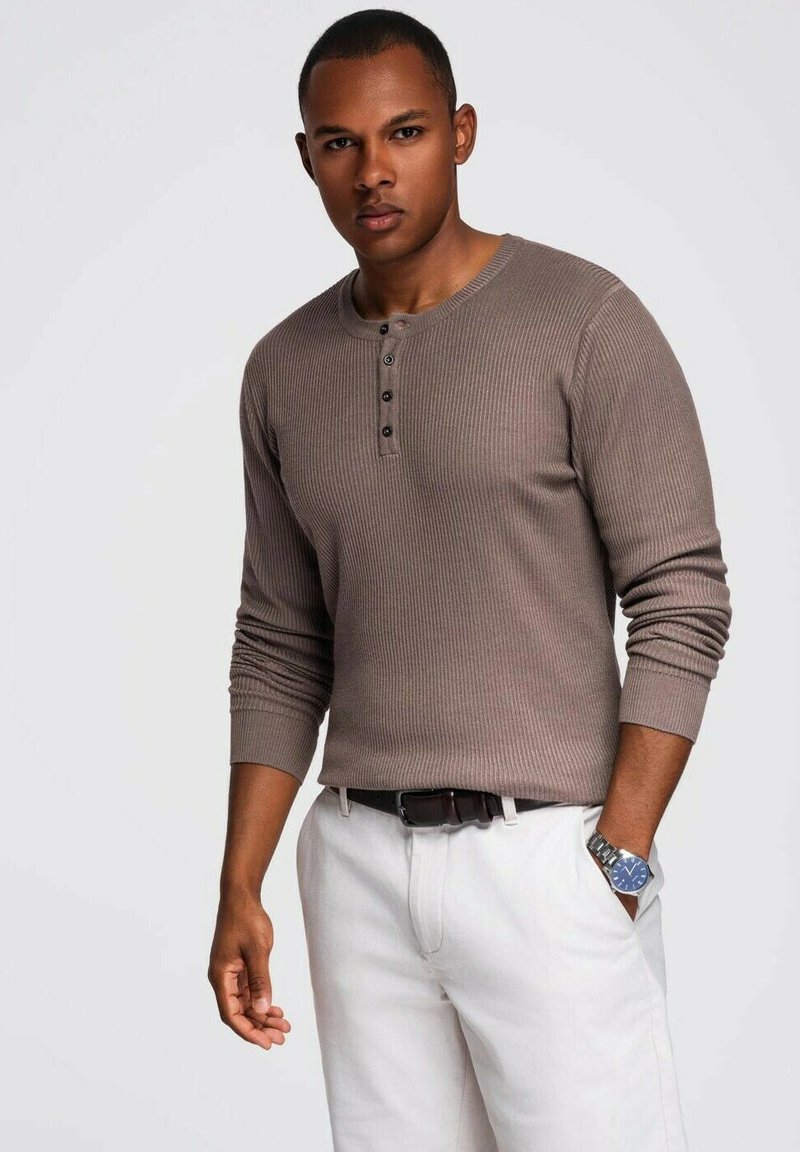 Camisa henley marrón de manga larga y canalé con tres botones negros en el cuello, combinada con pantalones claros. Se ve un reloj plateado en la muñeca.