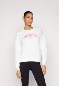 Sweatshirt blanc avec le texte rose "LA CONFIANCE SE JOUER SEULEMENT", avec un col rond, des manches longues et des poignets côtelés. Porté avec des leggings noirs.