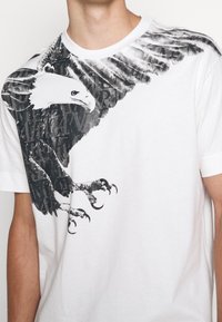 Bílý bavlněný tričko s černobílou grafikou orla se zachycenými detaily peří a drápů, částečně překrývající stylizovaný text.