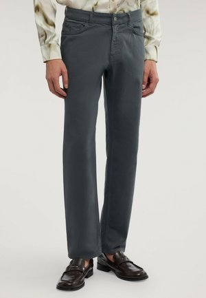 Man som bär mörkgrå raka byxor, bruna läderloafers och en ljus långärmad skjorta med ett diskret mönster.