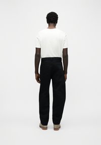 Άνδρας που στέκεται πλάτη φορώντας απλό λευκό μπλουζάκι, μαύρο τζιν και ανοιχτόχρωμα μποτάκια μπροστά σε απλό λευκό φόντο.