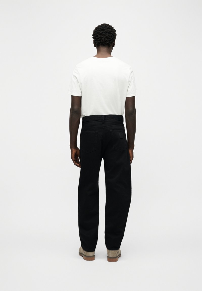 Mann steht mit dem Rücken zur Kamera, trägt ein schlichtes weißes T-Shirt, schwarze Jeans und helle Stiefel vor einem einfarbigen weißen Hintergrund.