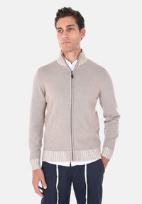Cardigan beige in maglia con zip, colletto e orlo a coste, caratterizzato da una trama uniforme e maniche lunghe, indossato sopra una camicia bianca.