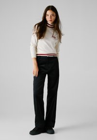 Weißer Pullover mit roten Streifen am Kragen und Saum, mit gesticktem Text. Schwarze weit geschnittene Hose und schwarze Schuhe runden das Outfit ab.