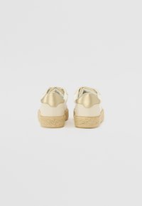 Bijele platnene espadrile s zlatnim detaljima, pletena jute potplat, i zaobljenim vrhom; stil koji se lako obuva s presavijenim tekstilnim detaljem na peti.
