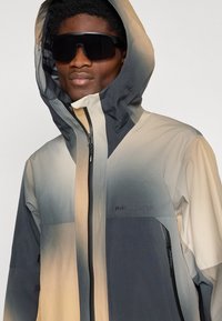Man wearing gradient beige and gray hooded jacket with front zip and black sunglasses against plain background.