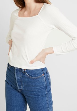 Frau trägt ein weißes, geripptes Langarmshirt mit quadratischem Ausschnitt und blaue, hochtaillierte Jeans, die Hände in die Hüften gestützt vor einem schlichten Hintergrund.