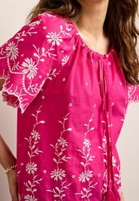 Fuchsia Baumwollbluse mit weißen Blumenstickereien, gerafftem Ausschnitt und Quasten-Detail. Kurze Ärmel mit gewelltem Saum.