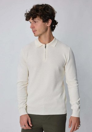 Homme aux cheveux bouclés portant un pull polo crème texturé à manches longues avec une fermeture éclair devant, debout devant un fond uni.