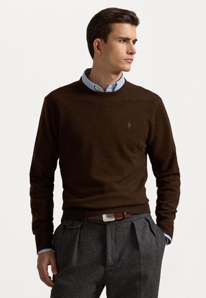 Bruine gebreide trui met een ronde hals, voorzien van een klein logo op de borst. Gecombineerd met grijze geplooide broek en een bruine riem met een metalen sluiting.