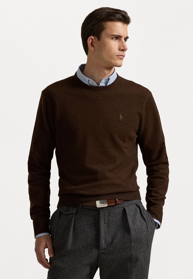 Suéter de punto marrón con cuello redondo, que presenta un pequeño logo en el pecho. Combinado con pantalones grises con pliegues y un cinturón marrón con un cierre de metal.