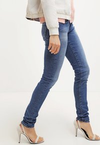 Slim-fit siniset farkut, joissa on sileä pinta, viisisalkkinen muotoilu ja huomaamattomat ompeluyksityiskohdat, yhdistettynä vaaleisiin korkeakorkoisiin kenkiin.