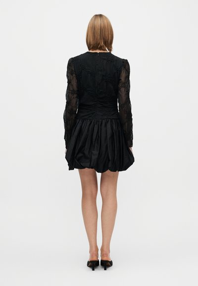 Rochie neagră cu mâneci lungi transparente, corset strâns pe corp și fustă evazată. Are broderii florale și închidere cu fermoar în spate.