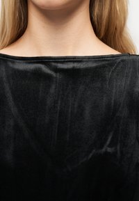 Κοντινό πλάνο του λαιμού και του άνω μέρους του στήθους ενός ατόμου που φοράει μαύρο βελούδινο τοπ χωρίς ώμους, με μακριά ξανθά μαλλιά ορατά.