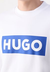 Biały sweter z niebieskim prostokątnym logo, na którym widnieje słowo "HUGO" napisane grubą białą czcionką. Ribnowany dekolt i gładka tekstura.
