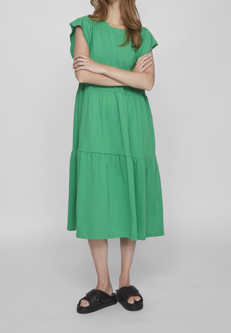 Femme portant une robe midi verte ample avec des manches à volants et des sandales compensées noires, bras croisés, debout sur un fond uni.