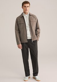 Homme debout les mains dans les poches, portant une veste taupe, un pull blanc à fermeture éclair quart, un pantalon sombre et des baskets blanches avec des semelles marron.
