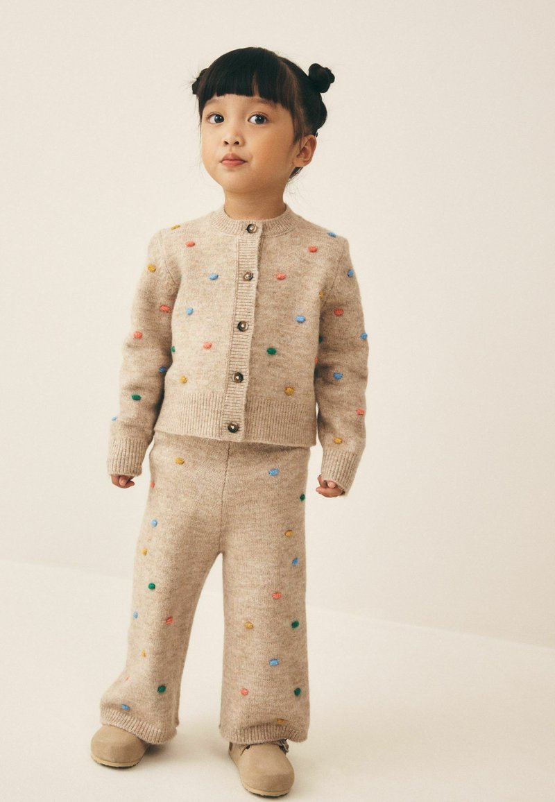 Young child with dark hair in two buns wearing beige knit sweater and pants with colorful pom-poms, standing on a light background.