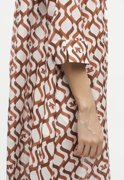 Χέρι και μπράτσο δίπλα σε μανίκι φορέματος με καφέ και λευκό κυματιστό μοτίβο και επαναλαμβανόμενα καφέ γράμματα "M" στον ύφασμα.