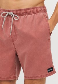 Burgundi színű fürdőnadrág, rugalmas derékkal, fehér zsinórral, oldalsó zsebekkel és fekete "RIP CURL" címkével a szegély közelében.