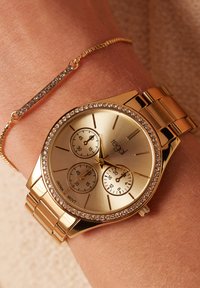 Gouden polshorloge met een ronde wijzerplaat, drie subdialen, kristallen accenten en een gouden metalen armband. Vergezeld van een delicate gouden kettingarmband.