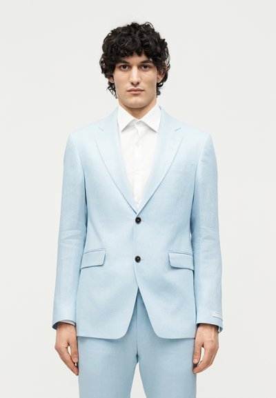 Άνδρας με σγουρά σκούρα μαλλιά, φορώντας ένα ανοιχτό μπλε σακάκι και παντελόνι πάνω από ένα λευκό κουμπωτό πουκάμισο, στέκεται σε απλό φόντο.