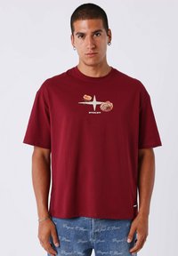 Joven vestido con una camiseta burdeos con un logotipo floral y de estrella, combinada con unos vaqueros azules que tienen un patrón de texto blanco por toda la prenda.