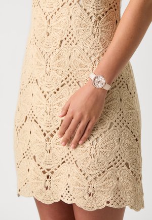 Abito in pizzo beige con motivo a farfalla; orologio da polso con quadrante rotondo, finiture in tonalità argento e cinturino chiaro.