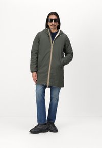 Olivenfarget, vanntett jakke med hette, hel glidelås med gule, hvite og blå striper, forlommer, sammen med blå jeans og svarte sko.