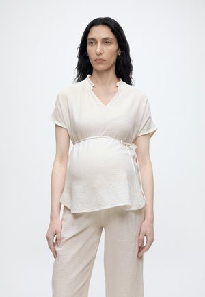 Kvinde med langt mørkt hår iført løs, kortærmet hvid top med sidesløjfe og matchende bukser med vide ben, stående mod en ensfarvet baggrund.