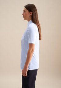 Camicia azzurra a maniche corte con colletto, taschino singolo sul petto, texture liscia e orlo dritto, indossata con pantaloni scuri.