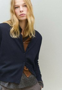 Námornícky modrý sveter s okrúhlym výstrihom a zapínaním na gombíky, položený na hnedom topu. Má jemnú textúru a priliehavé rukávy.