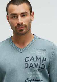 Hellblauer Strickpullover mit V-Ausschnitt, der gedruckten Text und Grafiken in Dunkelblau zeigt. Der Stoff wirkt strukturiert und lässig.