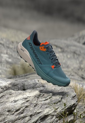 Chaussure de trail running Dynafit bleu et orange flottant au-dessus d'un terrain rocheux avec des plantes et un arrière-plan naturel flou.