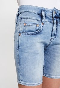 Jasnoniebieskie szorty dżinsowe mają dopasowany krój, pięć kieszeni i miedziane akcenty sprzętowe. Materiał ma gładką, lekko wyblakłą fakturę.